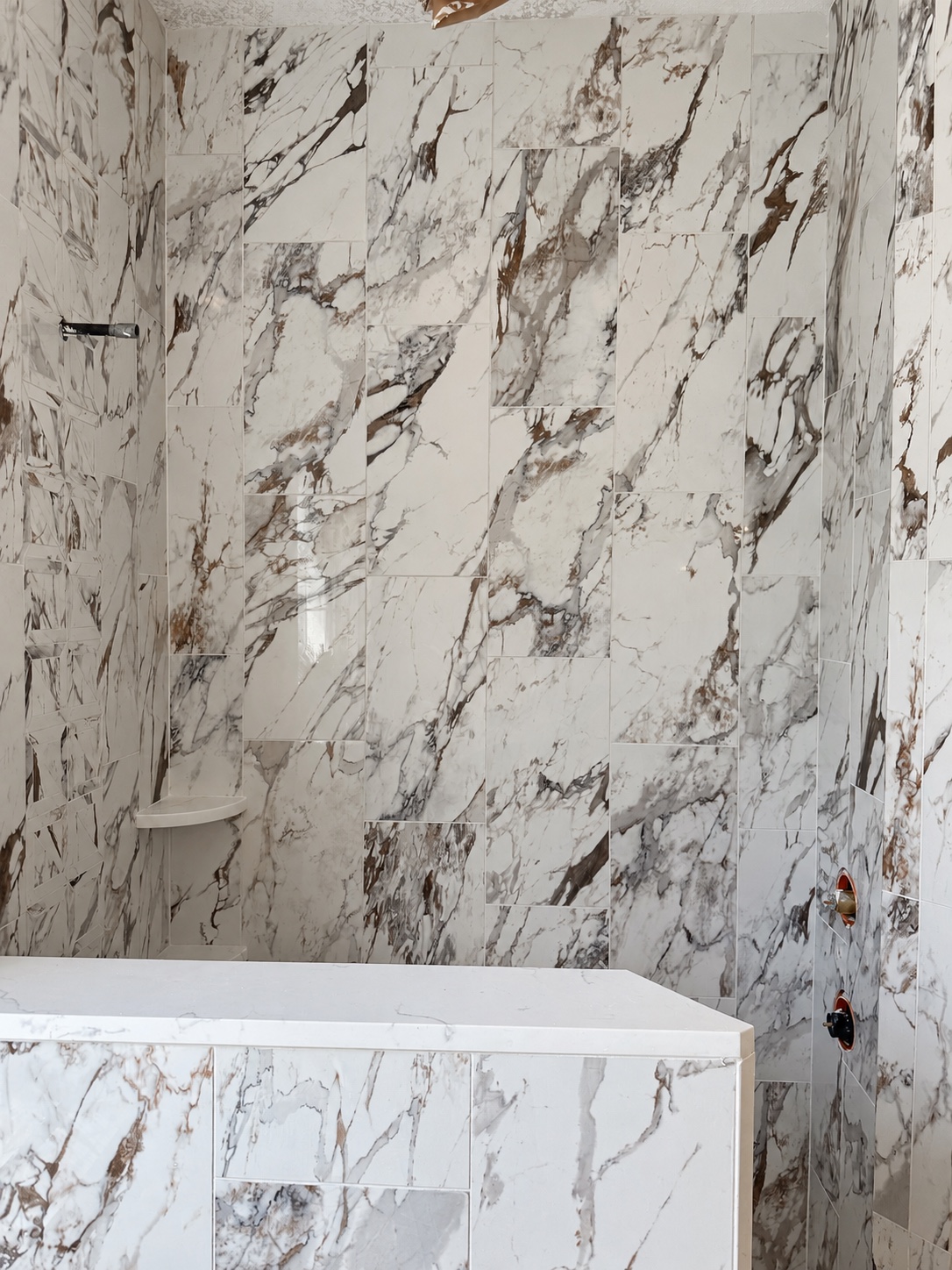 Warm marble-look shower surround with white corner shelves and a tiled half wall during installation