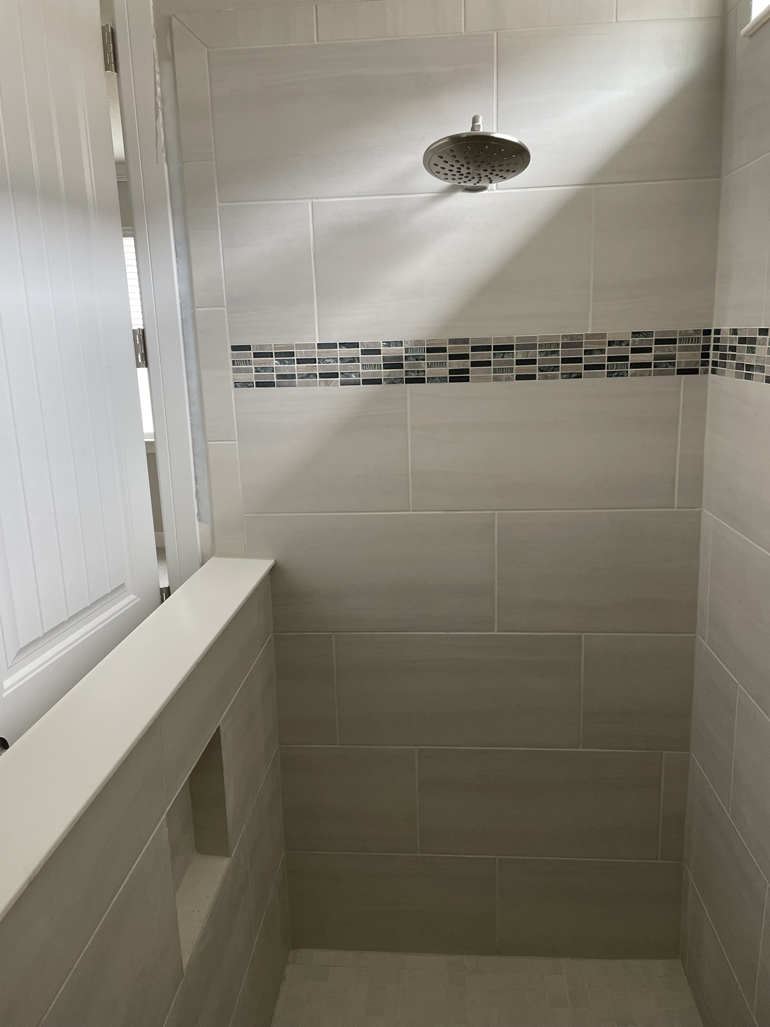 Light gray shower tile with a horizontal glass mosaic band and a rain shower head